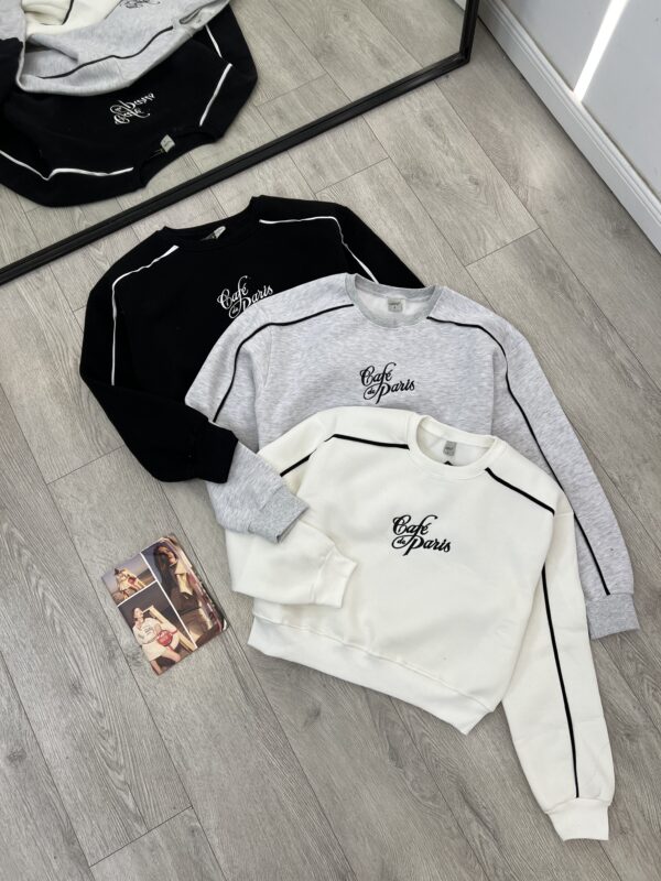 Bluza sportive me print "Café Paris" Ky imazh tregon tre bluza sportive me mëngë të gjata, të vendosura mbi një dysheme me parket. Ato kanë një dizajn të ngjashëm dhe janë në tre ngjyra të ndryshme.