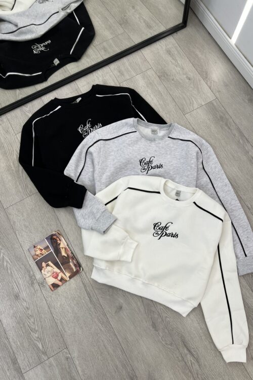 Bluza sportive me print “Café Paris” Ky imazh tregon tre bluza sportive me mëngë të gjata, të vendosura mbi një dysheme me parket. Ato kanë një dizajn të ngjashëm dhe janë në tre ngjyra të ndryshme.